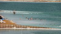 Landskabsbillede viser udsnit af stranden ved Det Døde Hav, hvor psoriasispatienter sendes i klimabehandling. En gruppe mennesker bader i det salte vand ved bredden og flyder. 